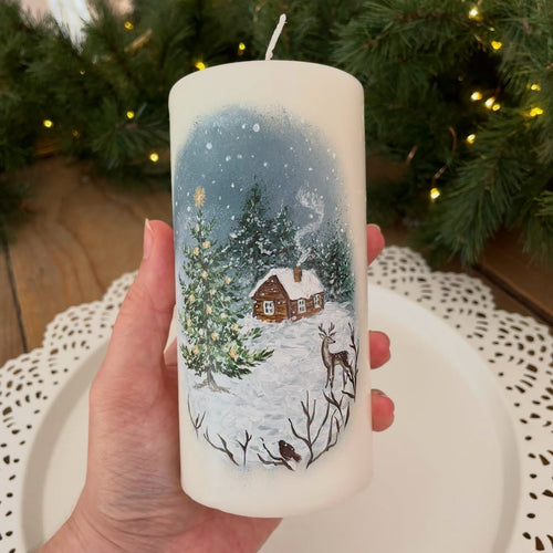 A hand holds a white cylindrical candle decorated with a hand-painted winter village scene featuring snow, pine trees, a small cabin, and a deer. The background shows a festive holiday setting with a blurred Christmas tree and warm lights.