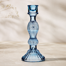 A light blue ribbed glass candlestick holder with a flared base, photographed on a neutral beige surface with a softly blurred background.