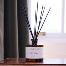 A reed diffuser bottle by Fox Cub & Co on a wooden table, with dark reeds extending upward against a softly blurred indoor background.