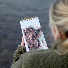 A person with blonde hair tied back, wearing a textured olive green fleece jacket, holds a spiral-bound notebook.