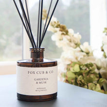 A reed diffuser bottle with black reeds, labeled 'FOX CUB & CO GARDENIA & MUSK', sitting on a dark tabletop in front of a softly blurred window and flowers.